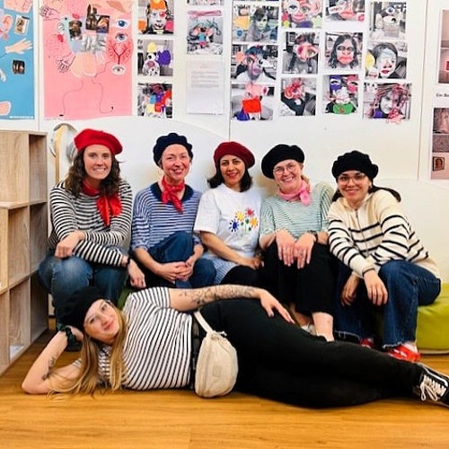 Das Team: Gruppe von Erwachsenen in einem Ausstellungsraum mit Kunstwerken an den Wänden, einige sitzen, andere liegen entspannt auf dem Boden.