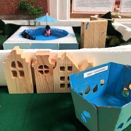 Modell einer Stadtlandschaft aus Holz, Karton und Naturmaterialien mit Häusern, Grünflächen und einem kleinen Schwimmbecken.
