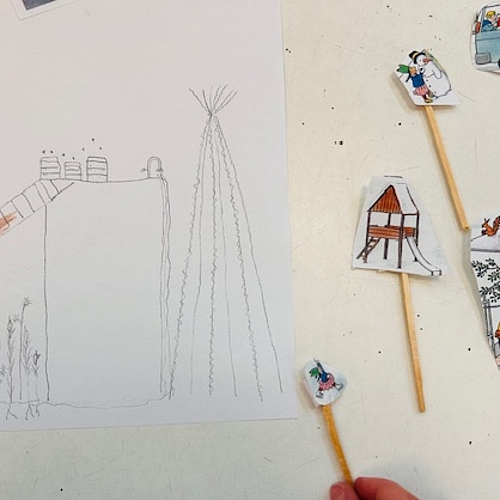 Auf einem Tisch liegt eine detailreiche Kinderzeichnung eines mehrstöckigen Hauses mit Rutsche, Pflanzen und einem Zelt im Garten. Daneben befinden sich kleine ausgeschnittene Papierfiguren auf Holzstäbchen, darunter ein Bus mit Kindern, ein Baumhaus, ein Café und einzelne Spielfiguren. Eine Hand hält eine der Figuren ins Bild. Die Szene zeigt kreative Auseinandersetzung mit Architektur und Lebensräumen.
