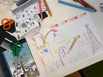 Auf einem Tisch liegt eine farbig gestaltete Kinderzeichnung eines mehrstöckigen Hauses mit Fenstern, Treppen, Pflanzen, Lampen und einer Rutsche. Daneben befinden sich Buntstifte, zwei Kinderscheren und ausgeschnittene Bilder aus einem Buch. Das Haus ist mit vielen Details und Farben versehen und zeigt kreative Ideen zu Architektur und Lebensräumen.
