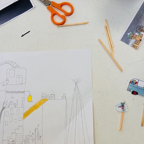 Auf einem Tisch liegt eine Kinderzeichnung eines fantasievollen Hauses mit mehreren Ebenen, Treppen, einer gelben Rutsche und kleinen Details wie Pflanzen und Fenstern. Daneben liegen ausgeschnittene Papierfiguren, Holzstäbchen, eine Kinderschere und Bastelmaterial. Das Bild zeigt kreative Entwürfe zum Thema Architektur und Nachhaltigkeit.