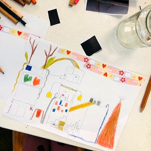Kinderzeichnung einer Stadt mit Häusern, Wegen, Pflanzen und farbigen Elementen auf einem Tisch mit Stiften.