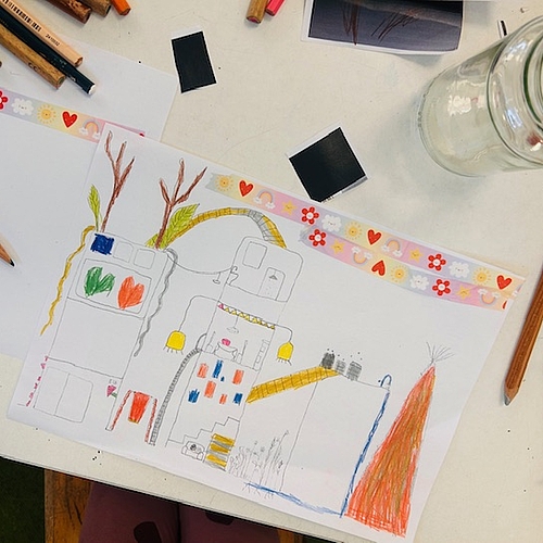 Auf einem Basteltisch liegt eine farbig ausgemalte Kinderzeichnung eines mehrstöckigen Hauses mit Fenstern, Pflanzen auf dem Dach, einer gelben Rutsche und einem angrenzenden Gartenbereich. Oben ist ein dekorativer Streifen mit Herzen und Blumen aufgeklebt. Rundherum liegen Buntstifte, ein Glasgefäß und Bastelmaterial. Die Zeichnung zeigt fantasievolle Ideen zu einem lebendigen, nachhaltigen Lebensraum.