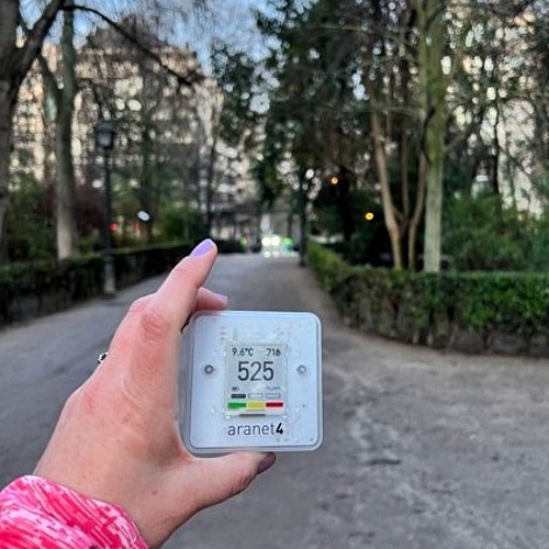 Hand hält ein digitales CO₂-Messgerät in einer begrünten Parkanlage mit Weg in Madrid und Bäumen im Hintergrund.